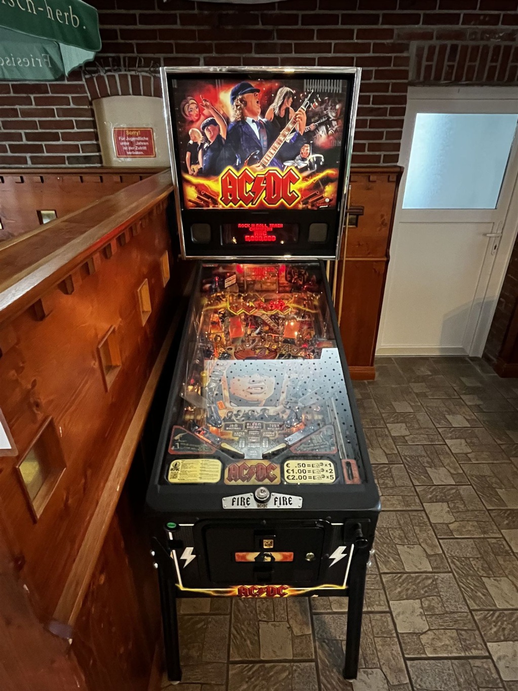 Ein Retro-Arcade-Spielgerät mit AC/DC-Motiv steht auf einem Holzbarhocker vor einer Ziegelwand.
