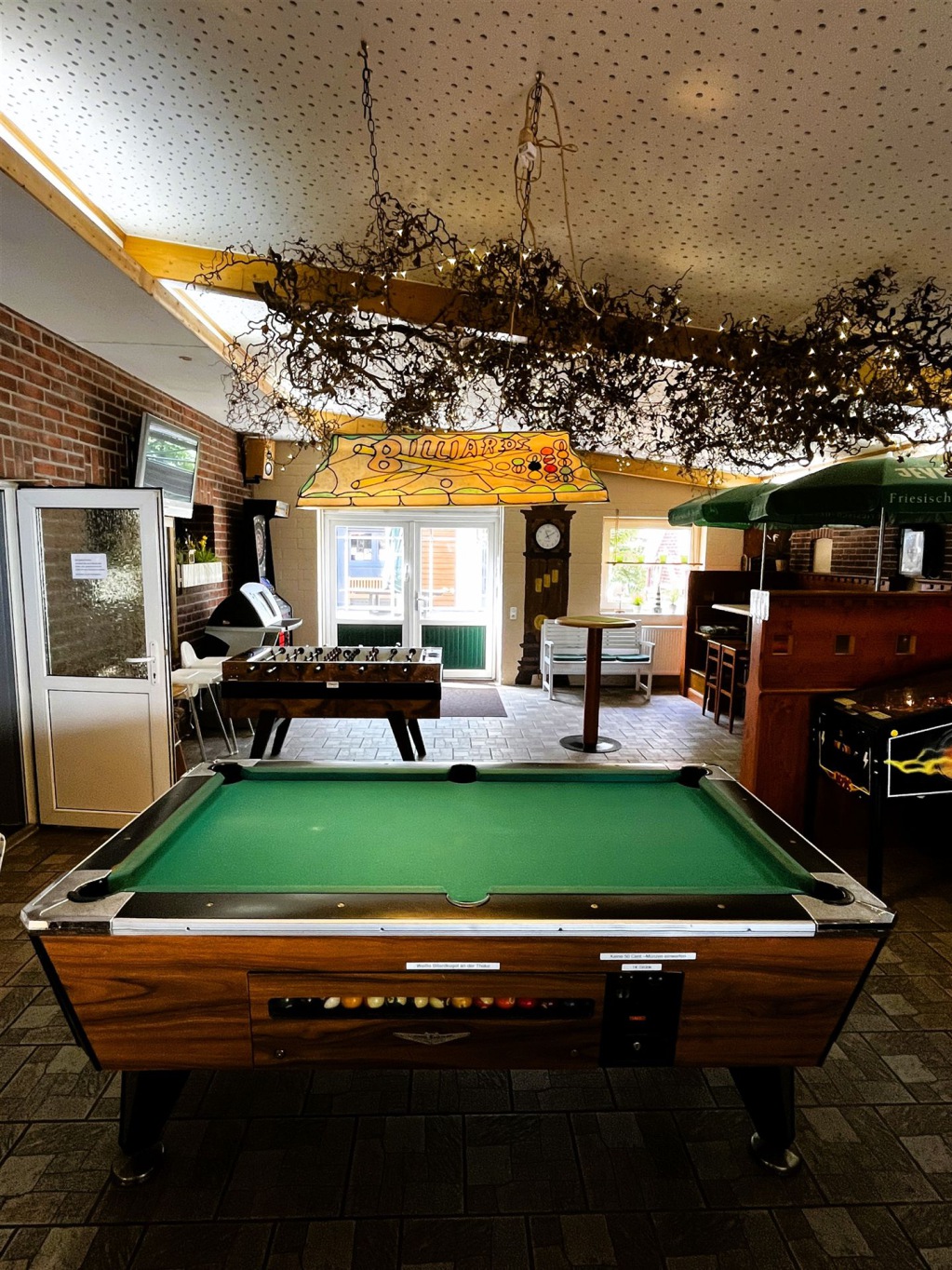 Ein großer Pooltisch mit grünem Tuch steht in einem gut beleuchteten Gastraum, der von dekorativen Ranken belebt wird.
